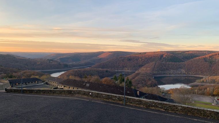 Eine malerische Landschaft mit sanften Hügeln und ruhigen Gewässern. Der Himmel erstrahlt in warmen Farben des Sonnenuntergangs.