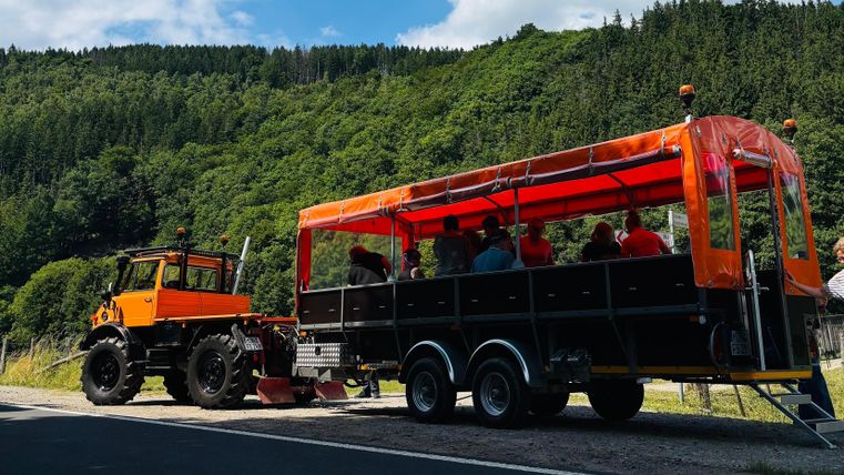 Ein Traktor zieht einen Wagen mit vielen Passagieren. Die Aussicht ist grün und bergig, mit einem blauen Himmel im Hintergrund.