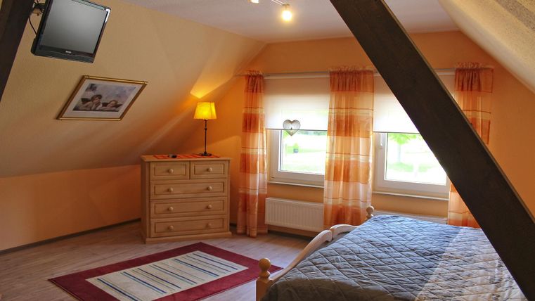 Ein gemütliches Schlafzimmer mit einem Doppelbett, einem Fernseher und einem Holzschrank. Große Fenster mit hellen Vorhängen sorgen für viel Tageslicht.