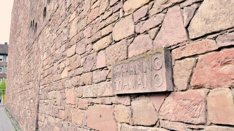 Eine historische Steinmauer mit einer beschrifteten Plakette. Die Mauer ist alt und zeigt typische Baustellenmerkmale.
