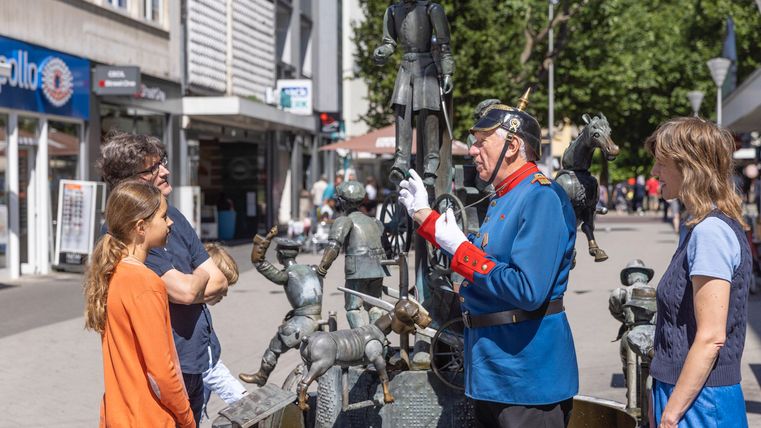 Eine Gruppe von Menschen steht um einen Brunnenskulptur in der Stadt. Ein Mann in Historienkleidung spricht mit zwei Kindern und einer Frau.