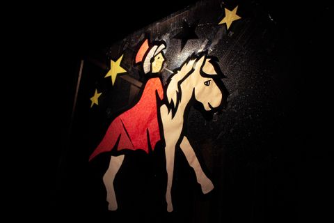 Eine bunte Papierfigur einer Reiterin in einem roten Kleid, die ein Pferd führt. Hintergrund ist dunkel mit goldenen Sternen.