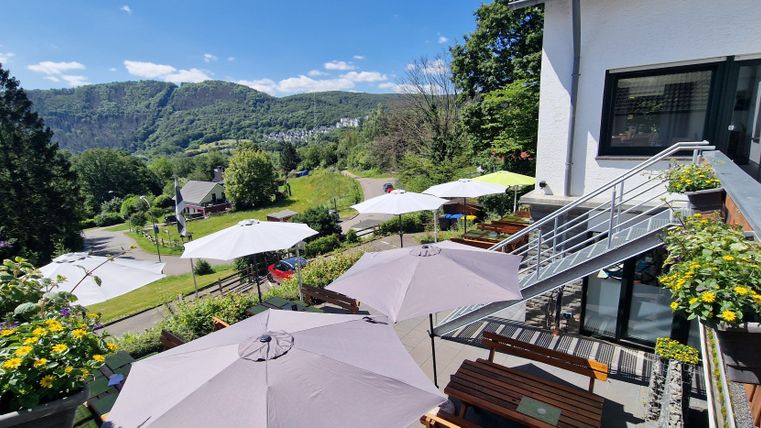 Eine sonnige Terrasse mit Sonnenschirmen und blühenden Pflanzen. Im Hintergrund sieht man eine grüne Landschaft mit sanften Hügeln.