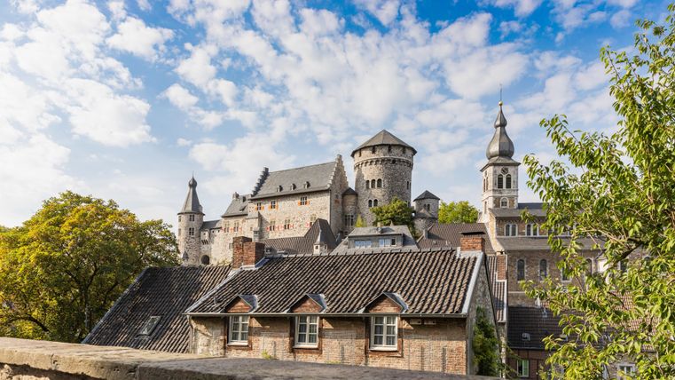 Eine beeindruckende Burg mit Türmen und Zinnen, umgeben von Bäumen. Der Himmel ist blau mit einigen Wolken.
