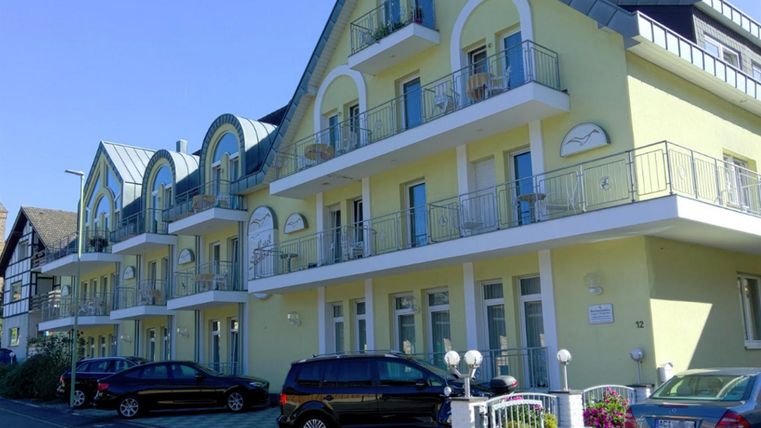 Ein modernes, gelbes Gebäude mit mehreren Balkonen. Autos parken vor der Fassade, und der Himmel ist blau.