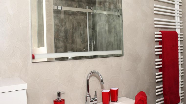 Ein modernes Badezimmer mit einem weißen Waschbecken und einem großen Spiegel. Rote Handtücher und Zubehör setzen einen Farbakzent auf das schlichte Design.