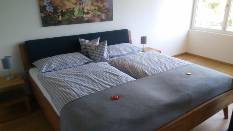 Ein gemütliches Schlafzimmer mit einem großen Doppelbett und grauer Bettdecke. An der Wand hängt ein Bild von lila Blumen und Fenster sorgen für viel Tageslicht.