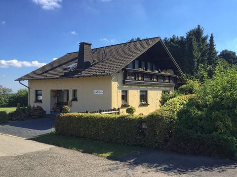 Ein gemütliches Haus mit einem Balkon und einem gepflegten Garten. Im Hintergrund sind Bäume und ein klarer Himmel zu sehen.