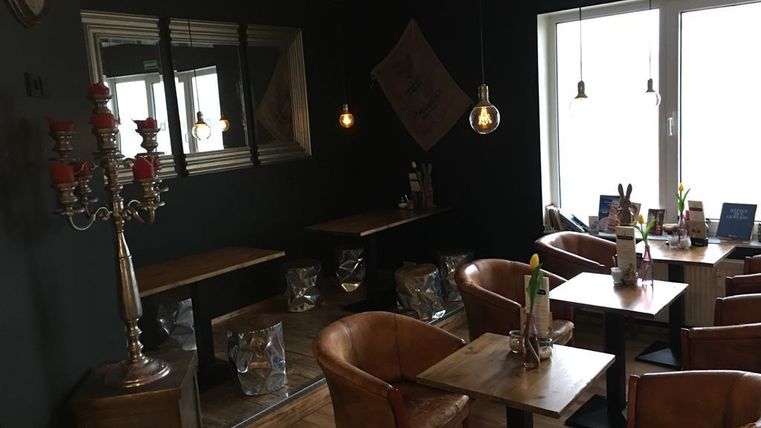 Ein gemütliches Café mit dunklen Wänden und Holztischen. Die Sitzbereiche sind mit eleganten Stühlen und sanfter Beleuchtung ausgestattet.