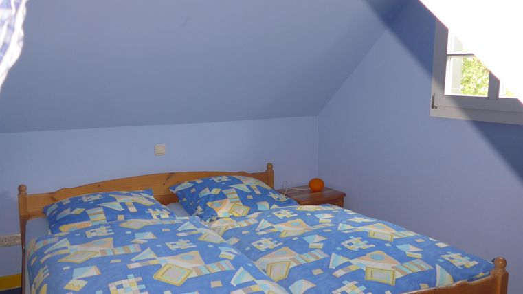 Ein gemütliches Schlafzimmer mit einem Doppelbett und blauer Wandfarbe. Das Bett hat eine bunte Bettwäsche und es gibt ein Fenster, das Tageslicht hereinlässt.