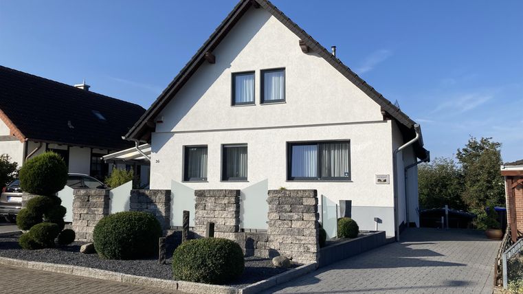 Ein modernes, zweigeschossiges Haus mit weißer Fassade und großen Fenstern. Der gepflegte Garten hat runde Sträucher und eine gepflasterte Einfahrt.