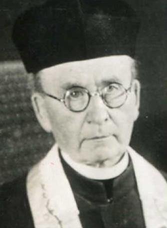 Ein älterer Mann mit Brille und schwarzer Kopfbedeckung. Er trägt eine dunkle Kleidung und hat einen ernsten Gesichtsausdruck.
