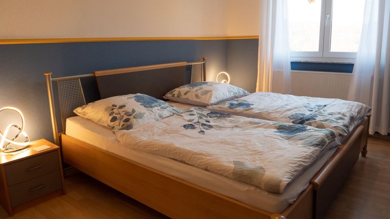 Ein gemütliches Schlafzimmer mit einem Doppelbett und hellen Bettdecken. Der Raum hat ein Fenster mit Vorhängen und einen Nachttisch mit einer Lampe.