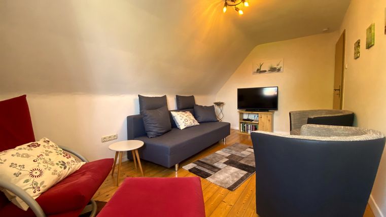 Ein gemütliches Wohnzimmer mit einer blauen Sofa-Garnitur und modernen Sesseln. Es gibt einen Fernseher und einen Holzboden, was eine einladende Atmosphäre schafft.