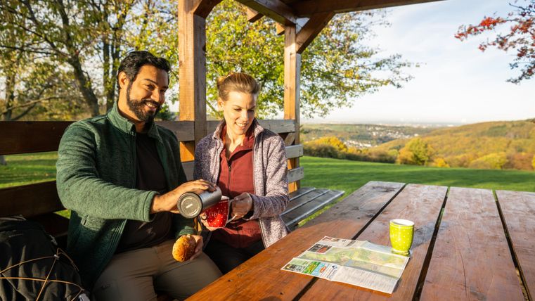 Ein Paar sitzt in einer Ruhezone unter einem Holzpavillon und genießt einen warmen Drink. Im Hintergrund sind Bäume und eine malerische Landschaft zu sehen.