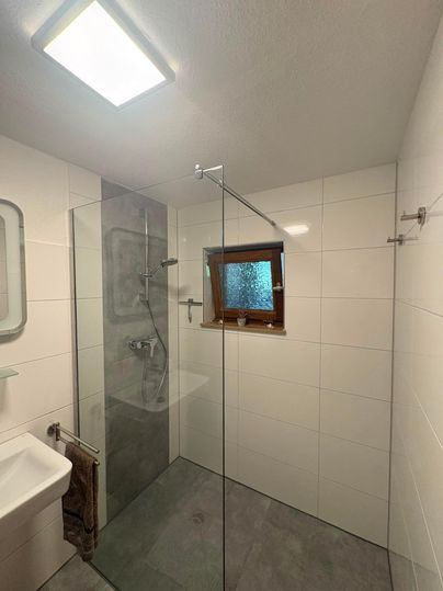 Ein modernes Badezimmer mit einer gläsernen Dusche und einem kleinen Fenster. Die Wände sind weiß und der Boden ist grau.
