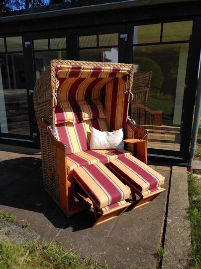 Ein gemütlicher Strandkorb mit gestreiften Kissen steht auf einer gepflasterten Fläche. Im Hintergrund sind große Fenster und ein grüner Garten zu sehen.