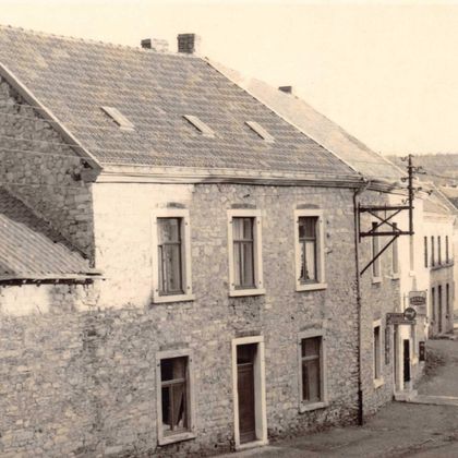 Ein altes, steinernes Haus an einer ruhigen Straße. Im Hintergrund sind mehrere weitere Gebäude und Bäume sichtbar.