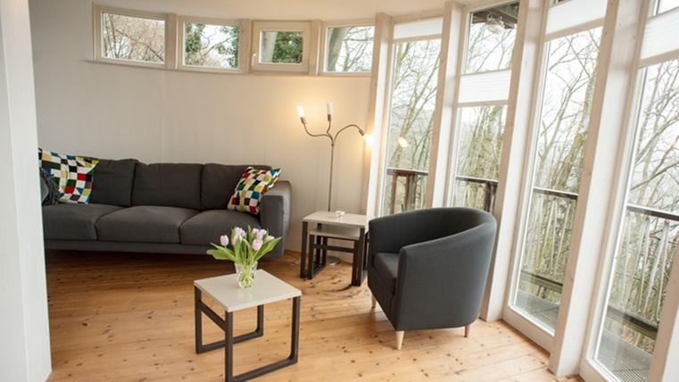 Ein gemütlicher Wohnraum mit einem grauen Sofa und einem schwarzen Sessel. Auf den Tischen stehen frische Blumen und große Fenster lassen viel Licht hereinfließen.