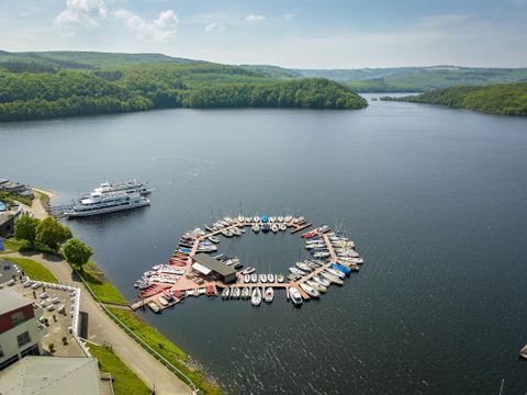 Eine idyllische Landschaft mit einem malerischen Hafen und vielen Booten auf einem ruhigen See. Im Hintergrund sind grüne Hügel und ein klarer Himmel zu sehen.