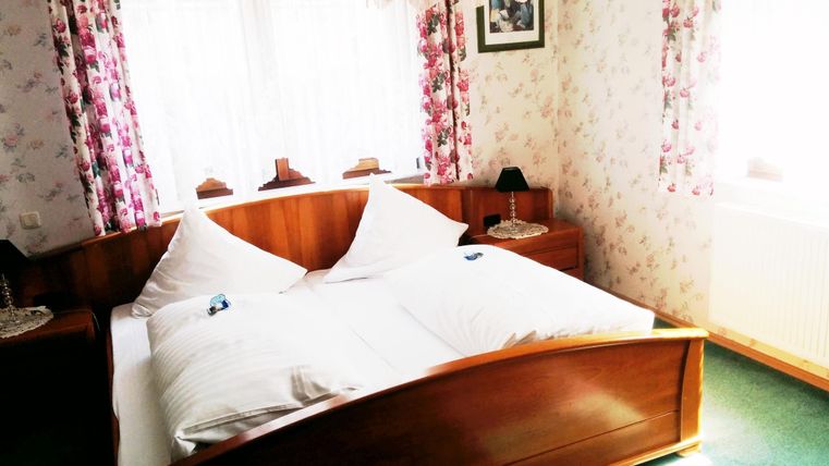 Ein gemütliches Schlafzimmer mit einem großen Bett und Blumenmuster an den Wänden. Helle Fenster sorgen für eine freundliche Atmosphäre.