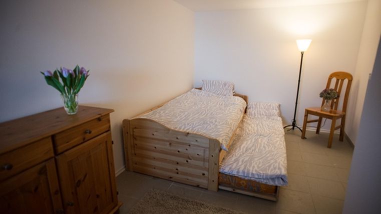 Ein einfaches Zimmer mit zwei Betten und einer Stehlampe. Auf einem Holzschrank stehen frische Blumen.