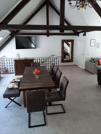 Ein moderner Essbereich unter einem hohen Dach mit sichtbaren Holzbalken. Der Tisch ist gedeckt und es gibt bequeme Stühle sowie ein gemütliches Sofa in der Ecke.