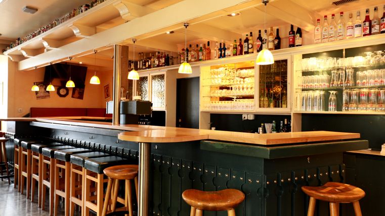 Eine moderne Bar mit Holzstühlen und einer langen Theke. Die Regale sind mit verschiedenen Getränken und Gläsern gefüllt.