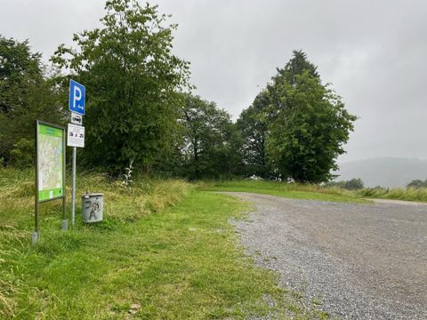 Ein Parkplatz in der Natur, umgeben von Bäumen und Gras. Der Himmel ist bewölkt, und der Weg führt ins Grüne.