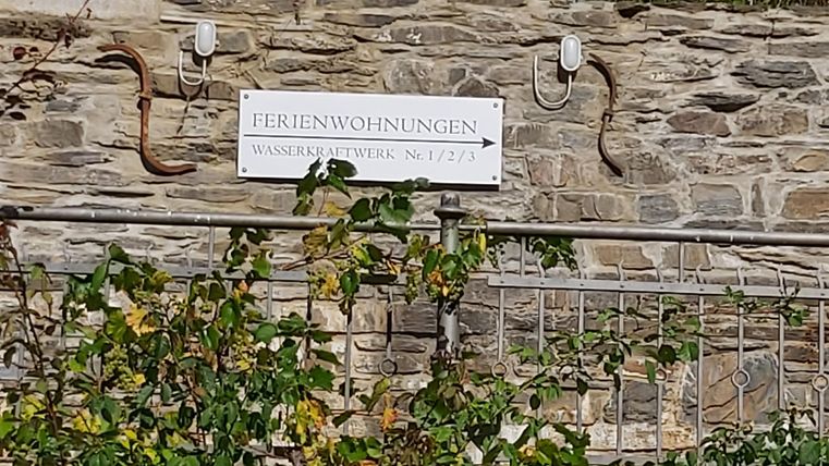 Ein Schild mit der Aufschrift "Ferienwohnungen" an einer Steinmauer. Es gibt Pflanzen und eine Richtungspfeil, der auf eine Unterkunft hinweist.
