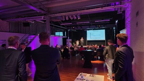 Eine Konferenz mit Teilnehmern, die einem Vortragenden auf der Bühne zuhören. Im Hintergrund sind Präsentationsfolien auf einem großen Bildschirm zu sehen.