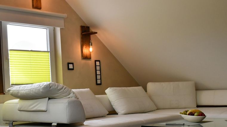Ein modernes Wohnzimmer mit einer weißen Couch und einem gläsernen Tisch. Natürliches Licht fällt durch das Fenster mit gelben Jalousien.