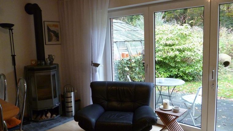 Ein gemütlicher Wohnraum mit einem schwarzen Sessel und einem Holzofen. Durch die Fenster blickt man in einen grünen Garten.