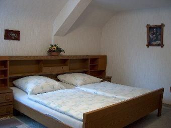 Ein einfaches Schlafzimmer mit einem Doppelbett und zwei Kissen. An der Wand hängen Bilder, und eine kleine Pflanze steht auf dem Nachttisch.