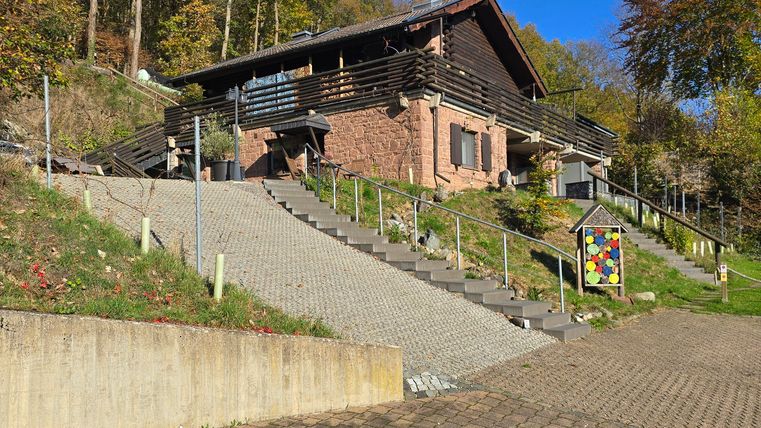 Ein gemütliches Haus auf einem Hügel, umgeben von Bäumen. Der blaue Himmel und die sonnige Umgebung verleihen der Szene einen freundlichen Eindruck.