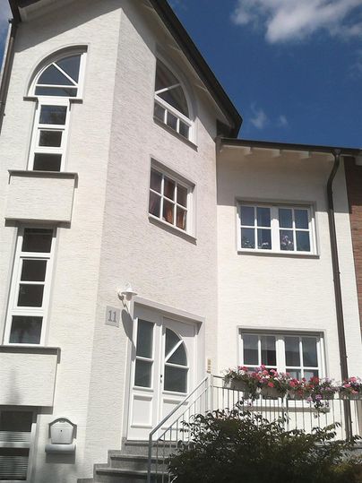 Ein modernes, weißes Wohnhaus mit großen Fenstern und einer geschwungenen Eingangstür. Im Vordergrund befindet sich eine Treppe mit einem Blumenbeet.