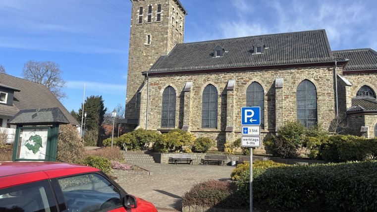 Eine alte Kirche mit einem markanten Turm steht im Vordergrund. Im Vordergrund ist ein rotes Auto und rundherum sind gepflegte Pflanzen und eine Parkzone zu sehen.