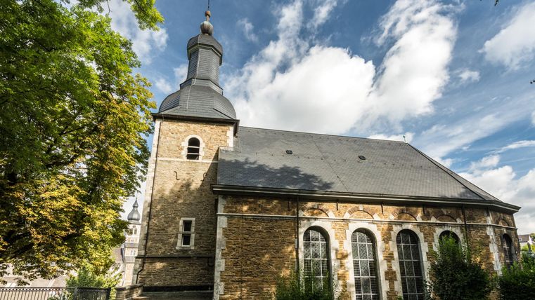 Das Bild zeigt eine kleine Kirche mit einem charakteristischen Turm und großen Fenstern. Umgeben von Grünflächen und Bäumen strahlt sie eine ruhige Atmosphäre aus.