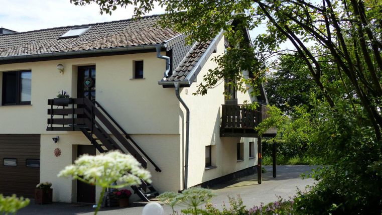 Ein modernes Haus mit einem Garten und einer Treppe zu einem Balkon. Umgeben von Bäumen und Blumen wirkt die Umgebung einladend und freundlich.
