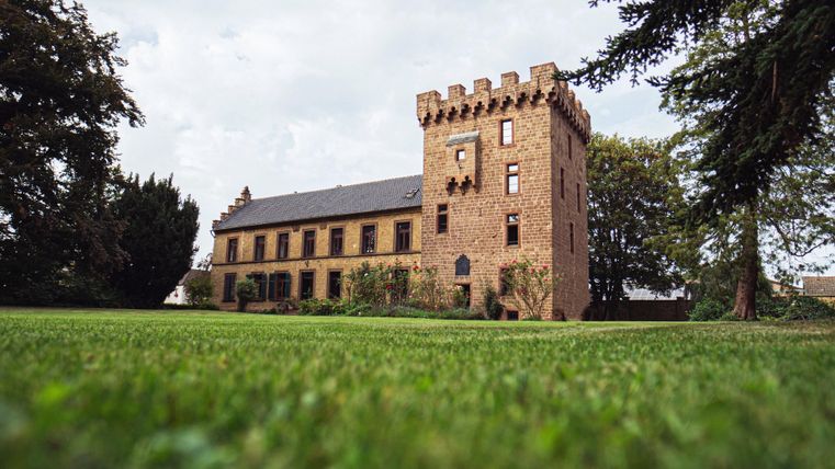 Ein historisches Gebäude mit einem quadratischen Turm und großen Fenstern. Der umgebende Garten ist grün und gepflegt.