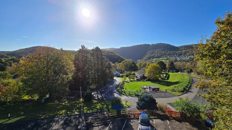 Eine malerische Landschaft mit grünen Bäumen und sanften Hügeln. Die Sonne scheint hell am blauen Himmel.