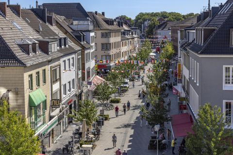 Eine belebte Einkaufsstraße mit Bäumen und Geschäften. Viele Menschen schlendern entlang der Straße.