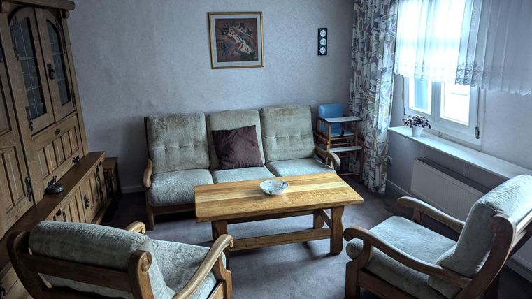 Ein gemütliches Wohnzimmer mit einer Holzbank, einem Tisch und einfachen Möbeln. Das Licht fällt sanft durch ein Fenster, und es gibt eine Wanddekoration.