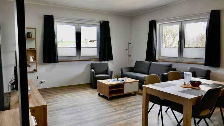 Ein modernes Wohnzimmer mit einem grauen Sofa, einem Couchtisch und einem Esstisch. Große Fenster lassen viel Licht herein und die Wände sind in neutralen Farben gehalten.