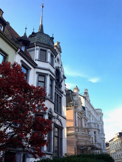 Eine beeindruckende Architektur mit historischen Gebäuden und einem strahlend blauen Himmel. Im Vordergrund ist ein roter Baum zu sehen.