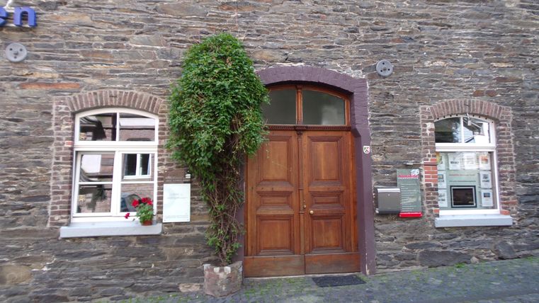 Eine alte Steinmauer mit einer großen Holztür in der Mitte. Links und rechts sind Fenster mit Blumen in den Fensterbänken.