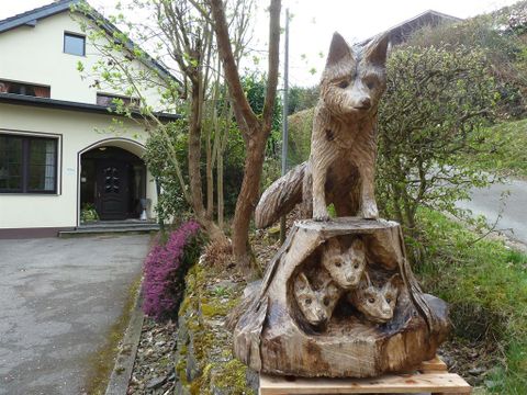 Eine Holzskulptur eines Fuchses und seiner Jungen steht vor einem Haus. Die Statue ist detailreich und zeigt die Tiere in einem natürlichen Umfeld.