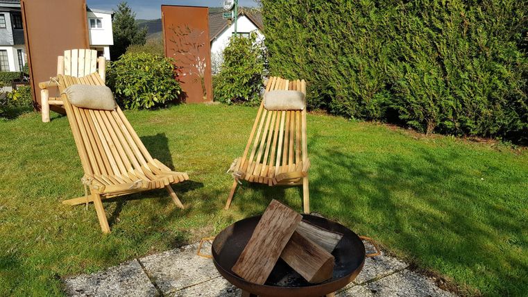 Ein schöner Garten mit zwei Holzliegestühlen und einem Feuerplatz. Die Umgebung ist grün und einladend.