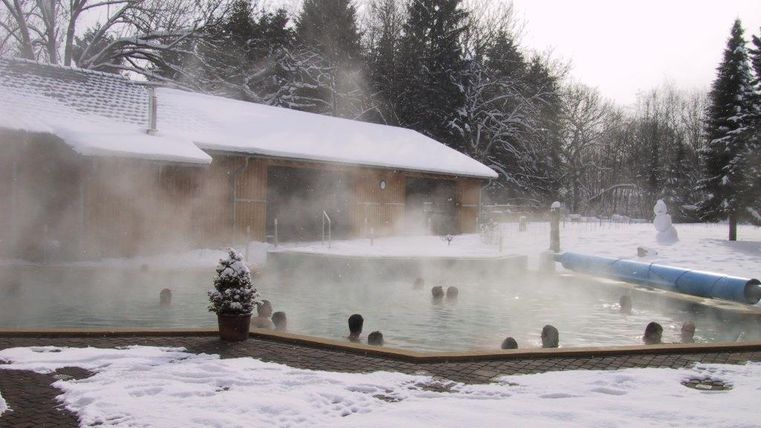 Ein beheiztes Schwimmbad im Freien, umgeben von Schnee und Dampf. Im Hintergrund steht ein Holzgebäude und ein paar Bäume sind sichtbar.