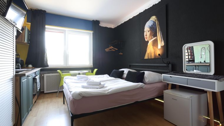 Ein modernes Zimmer mit einem Doppelbett und einem künstlerischen Wandbild. Es gibt einen Schreibtisch, einen Kühlschrank und viel Licht durch das Fenster.